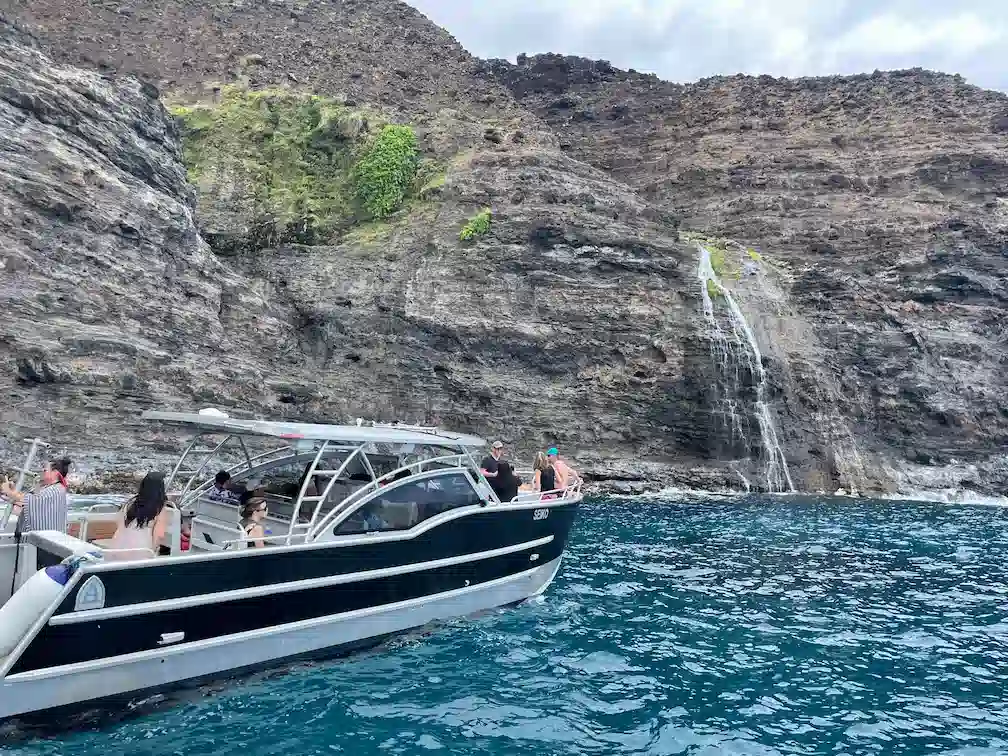 A Makana Charters tour boat floating in turquoise water beneath a natural waterfall cascading down the dramatic Na Pali Coast sea cliffs on a Na Pali Coast boat tour, Kauai, Hawaii.