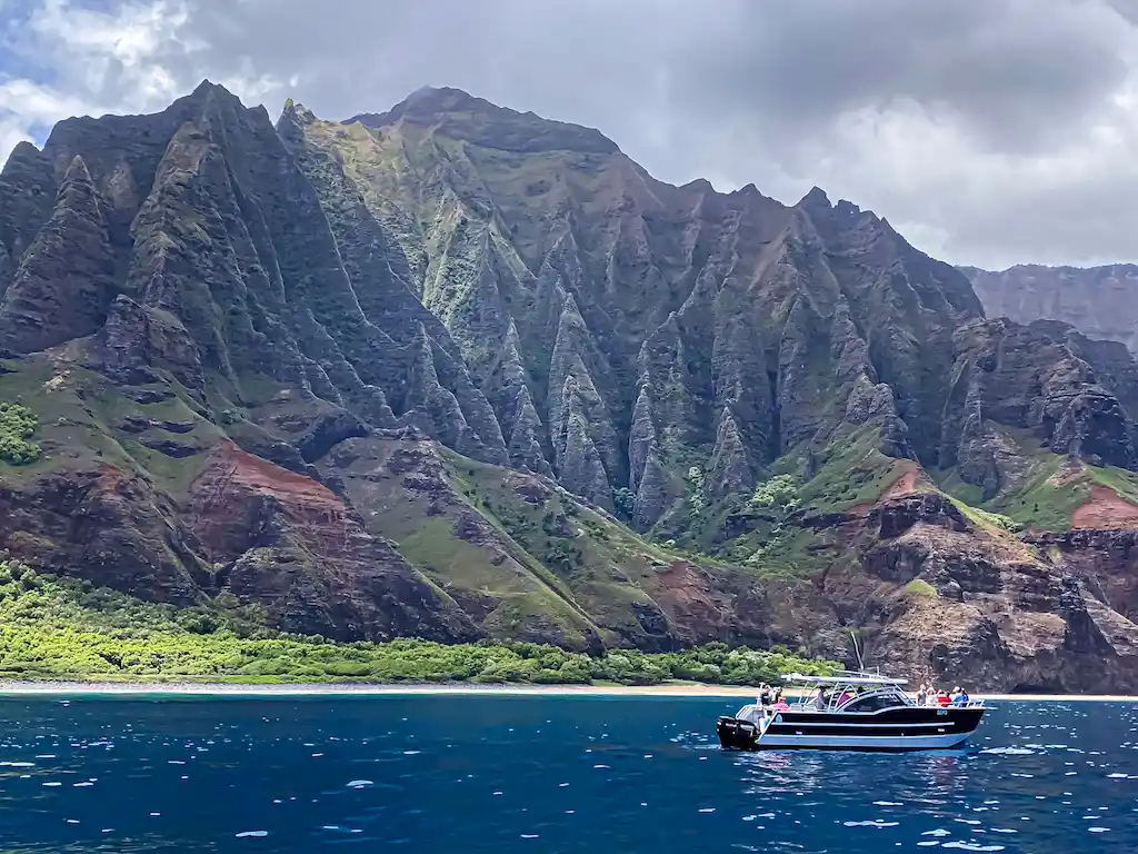 A Makana Charters tour boat cruising in turquoise water beneath the towering, cloud-shrouded green and dark Na Pali Coast sea cliffs on a Na Pali Coast boat tour, Kauai, Hawaii.
