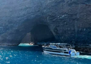 A Makana Charters tour boat floating in vivid turquoise water at the entrance of a dramatic Na Pali Coast sea cave with towering dark volcanic rock walls on a Kauai sea caves tour, Hawaii.
