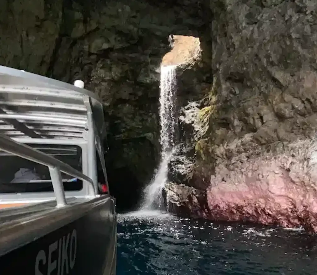 A Makana Charters tour boat alongside a cascading waterfall inside a dramatic Na Pali Coast sea cave with vivid blue water on a Kauai sea caves tour, Hawaii.
