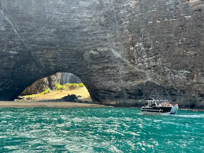 A Makana Charters tour boat floating in vivid turquoise water at the base of a massive Na Pali Coast sea cave arch with a sandy beach visible through the opening on a Kauai sea caves tour, Hawaii.