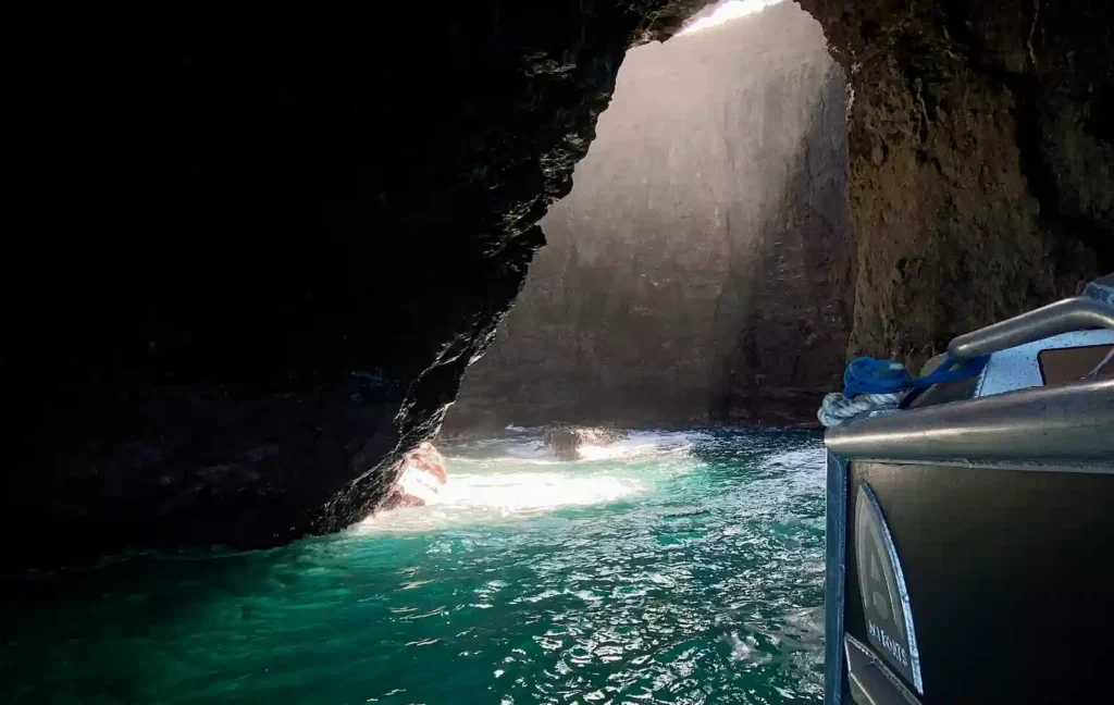 A dramatic shaft of light streaming into a dark Na Pali Coast sea cave illuminating the turquoise water and rocky walls beside a Makana Charters tour boat on a Kauai sea caves tour, Hawaii.