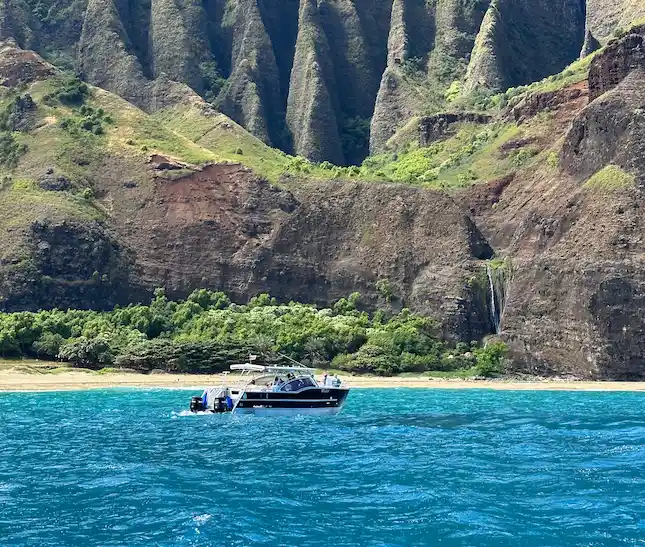 A Makana Charters tour boat anchored in calm turquoise water beneath the towering green and rust Na Pali Coast sea cliffs near a secluded beach on a Na Pali Coast boat tour, Kauai, Hawaii.