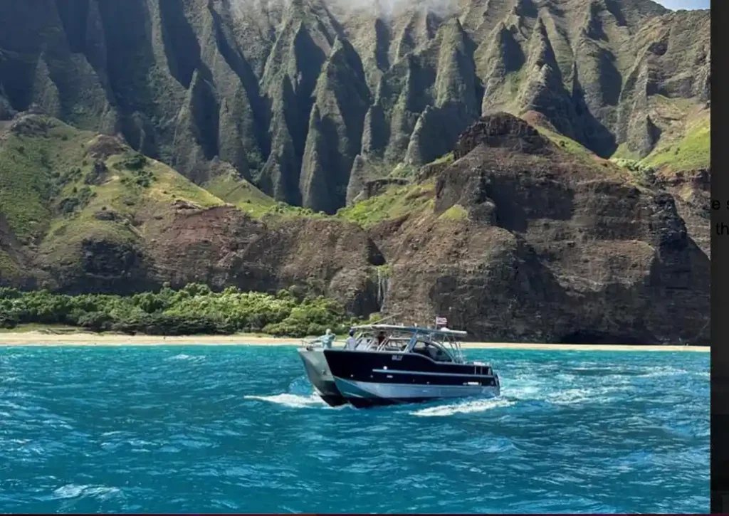 A sleek black Makana Charters tour boat speeding through turquoise water along the base of the lush green Na Pali Coast sea cliffs on a Na Pali Coast boat tour, Kauai, Hawaii.