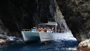 A white catamaran tour boat filled with passengers navigating through a narrow opening of a rocky sea cave.