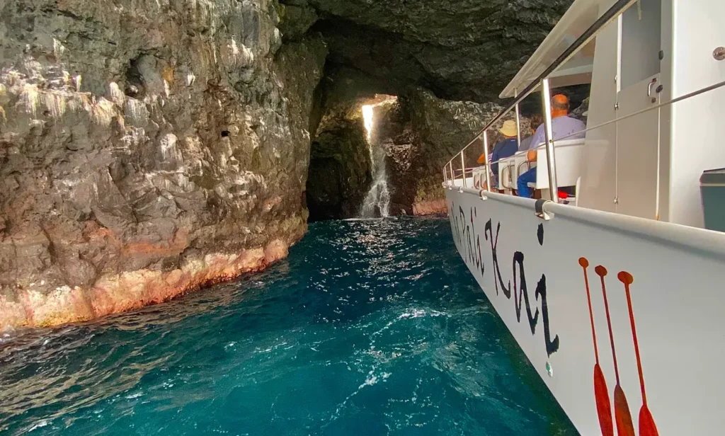 The Na Pali Kai tour boat navigating through a narrow Kauai sea cave with a waterfall trickling down the volcanic rock walls into vivid turquoise water on a Kauai sea caves tour.