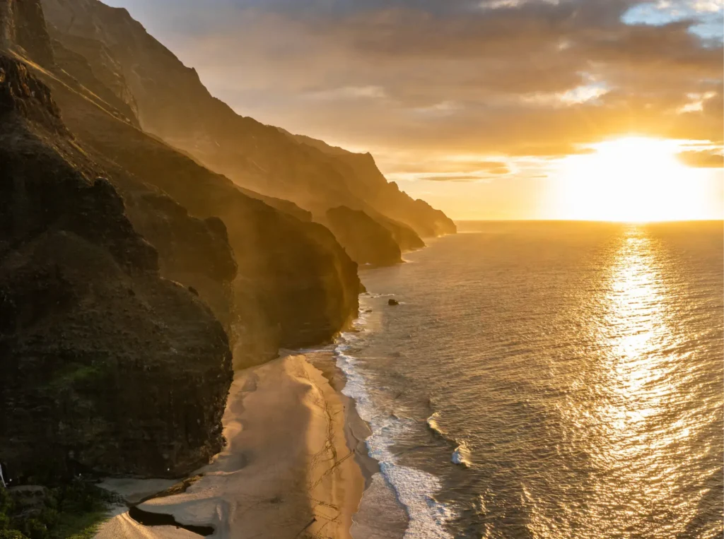 A breathtaking aerial view of a golden sunset over the Na Pali Coast's dramatic sea cliffs and secluded beach stretching along the Kauai coastline, Hawaii.