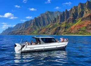 The Leialoha, a Makana Charters tour boat, cruising in vivid turquoise water beneath the towering green and rust Na Pali Coast sea cliffs on a Na Pali Coast boat tour, Kauai, Hawaii.