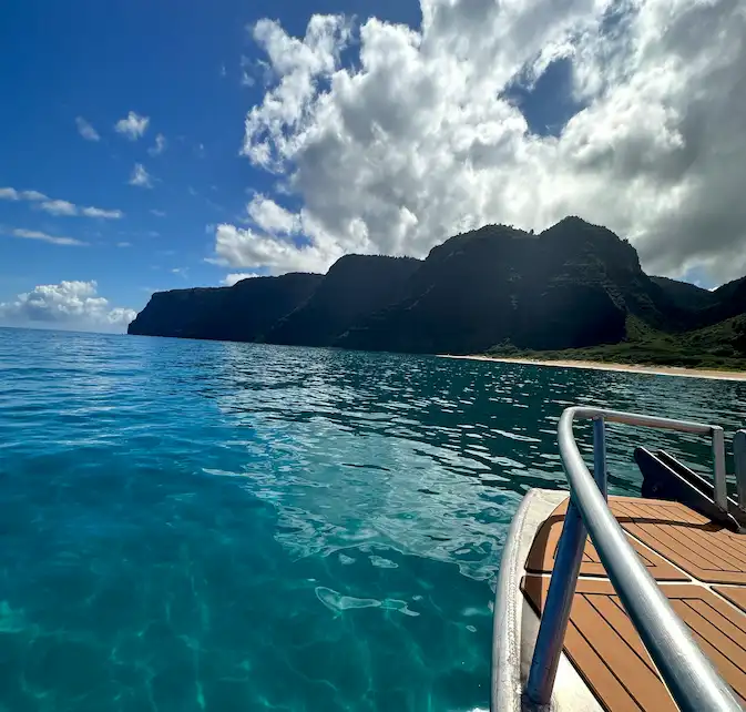 The bow of a Makana Charters tour boat gliding through mirror-calm turquoise water toward the dramatic Na Pali Coast sea cliffs under a bright blue sky with dramatic clouds, Kauai, Hawaii.