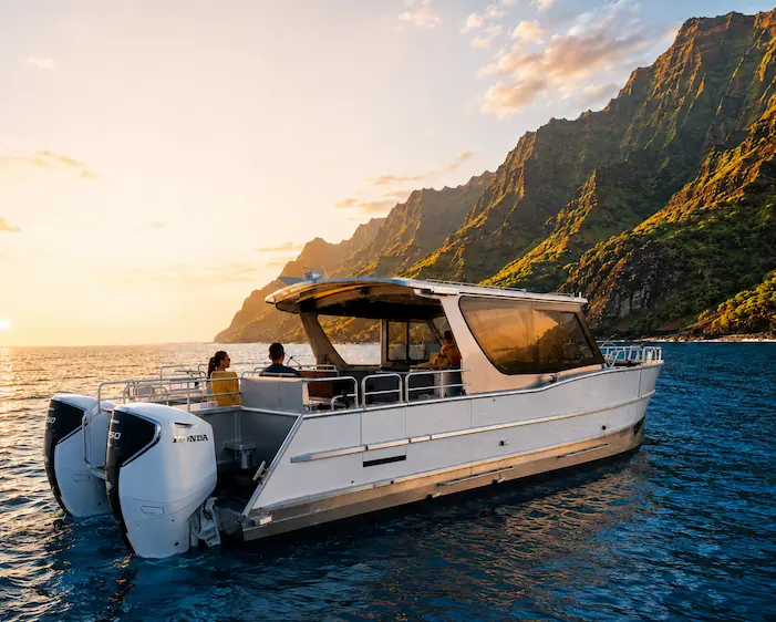The Amelia K, a Makana Charters luxury tour boat, anchored in calm golden water at sunset beneath the dramatic Na Pali Coast sea cliffs on a Na Pali Coast sunset cruise, Kauai, Hawaii.