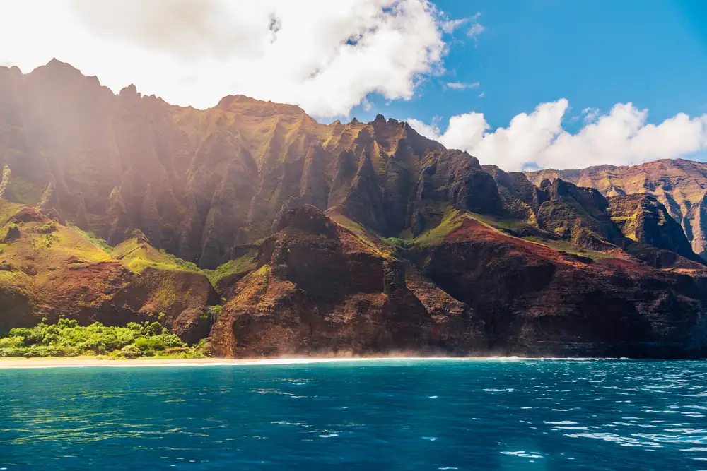 A vivid ocean-level view of the Na Pali Coast's sunlit green and rust-colored ridgelines and sea cliffs rising above the turquoise water on a Na Pali Coast boat tour, Kauai, Hawaii.