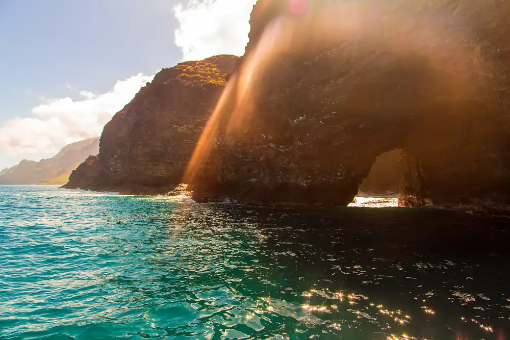 Golden sunlight streaming through a gap in the dramatic Na Pali Coast sea cliffs over vivid turquoise water on a Na Pali Coast boat tour, Kauai, Hawaii.