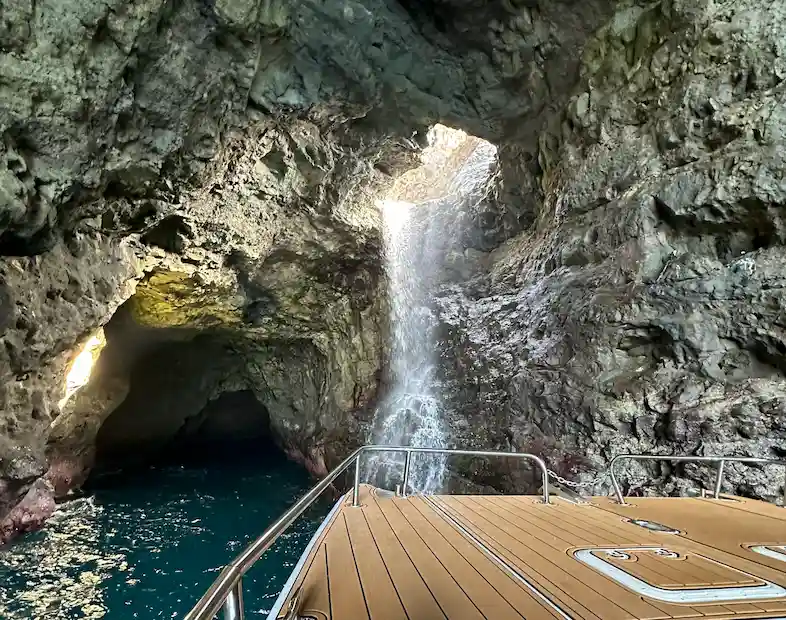 The deck of a Makana Charters tour boat inside a dramatic Kauai sea cave with a powerful waterfall cascading down the volcanic rock walls and natural light streaming through the cave opening above, on a Kauai sea caves tour, Na Pali Coast, Hawaii.