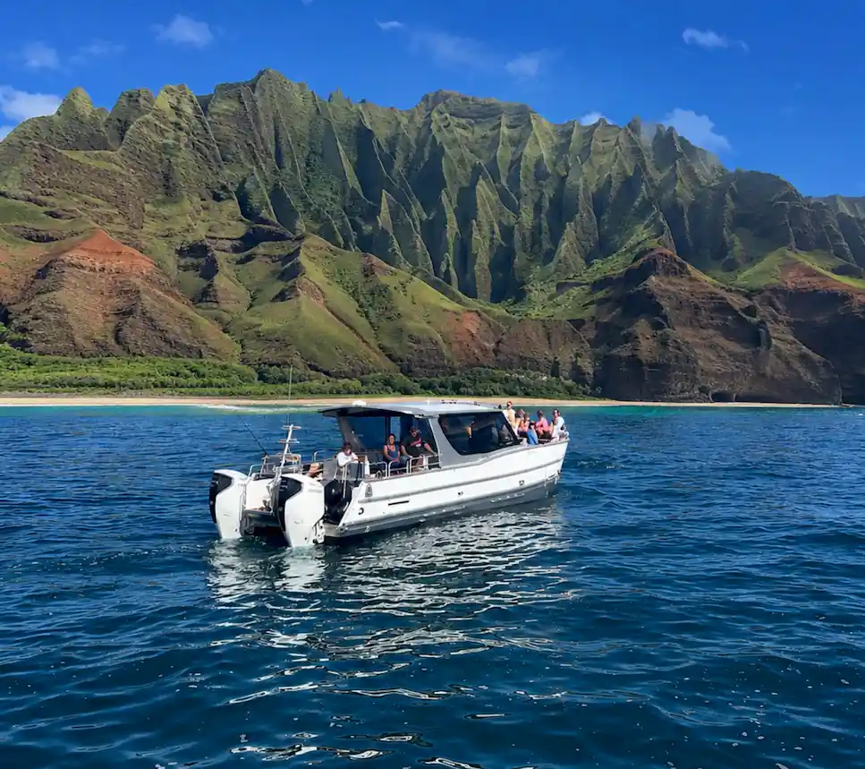 The Leialoha, a Makana Charters tour boat, anchored in calm turquoise water beneath the towering green and rust Na Pali Coast sea cliffs on a Na Pali Coast boat tour, Kauai, Hawaii.