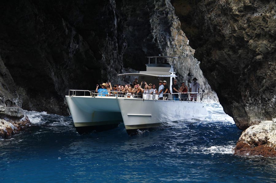A white catamaran tour boat filled with passengers navigating through a narrow opening of a rocky sea cave.